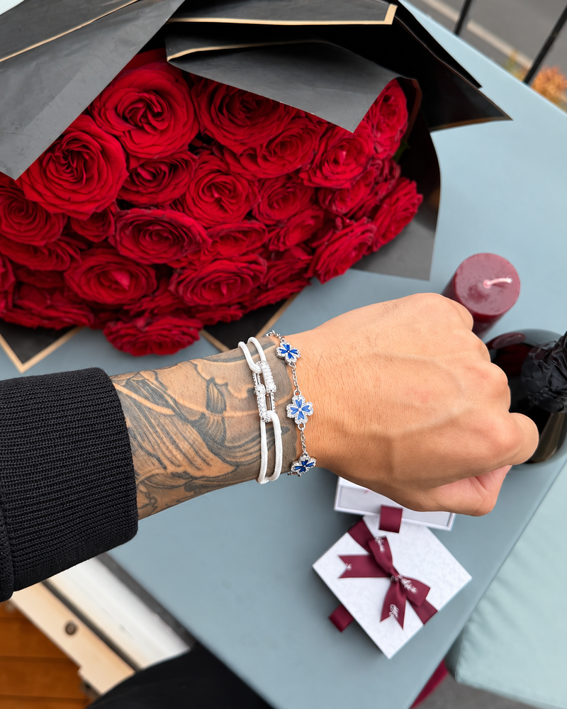 Koluré LIEN D’ÉCLAT Bracelet Blue – silberfarbenes Blütenarmband mit blauen Blütenflächen und weißem Stoffarmband vor roten Rosen